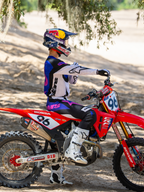 Motocrossförare som bär Limited Edition HUNTA96 XXV Supertech Vision Goggles i Purple White Mirror Silver, matchande lila och vit racingtröja med vita stövlar, sittande på en röd Honda motocrosscykel med nummer 96, utomhusmiljö för terrängkörning på en sandig bana.
