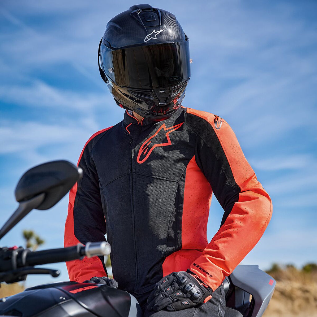Alpinestars Viper Air V4-jacka i röd fluo och svart, bärs av en förare på en motorcykel, med en stor logotyp på bröstet och ventilerade meshpaneler, kombinerad med en kolfiberhjälm och svarta handskar, väg- och sportmotorcykelutrustning
