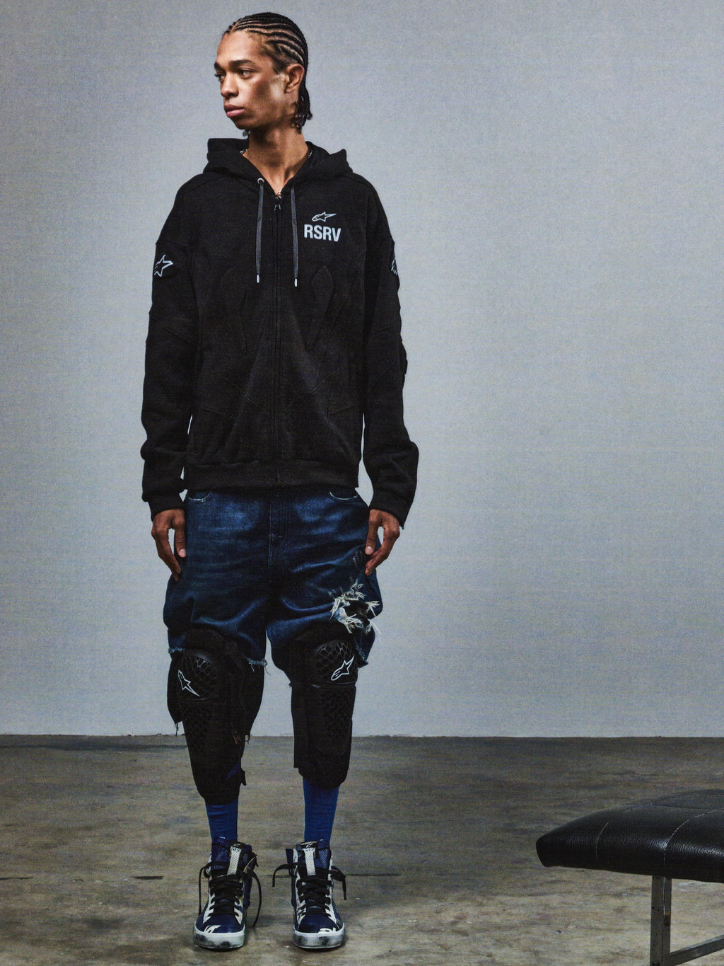 Alpinestars RSRV GP hoodie, svart premiumhuvtröja med RSRV-logotyp och stjärndetaljer, unisex streetwear från lyxkollektion, visad med slitna denimshorts och knäskydd