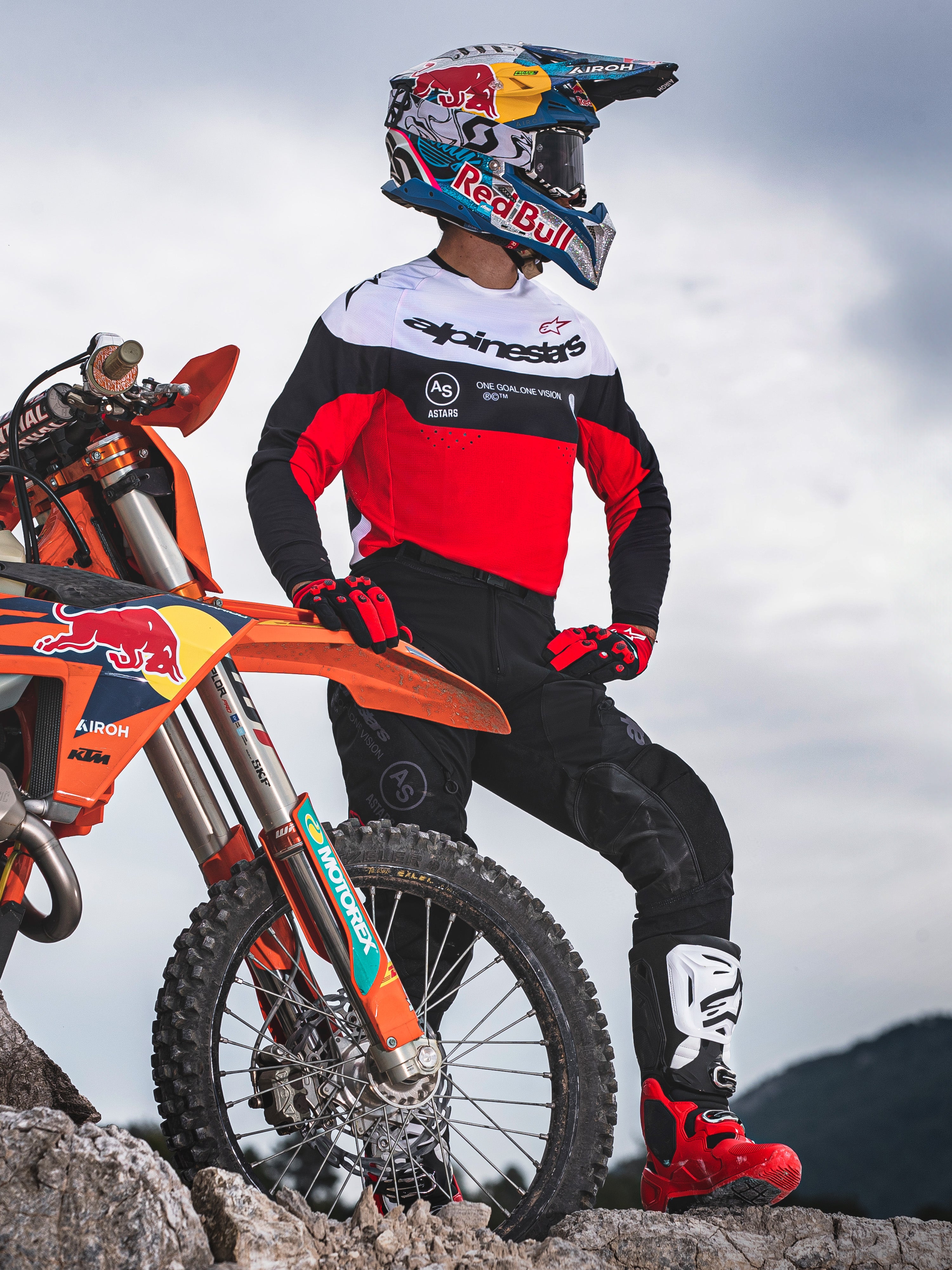 Motocrossförare iförd Alpinestars Pro-Dura-tröja i svart, ljusrött och vitt, stående bredvid en KTM-crosscykel i stenig terräng, matchad med svarta byxor och röda motocross-stövlar, utomhusmiljö för enduro.