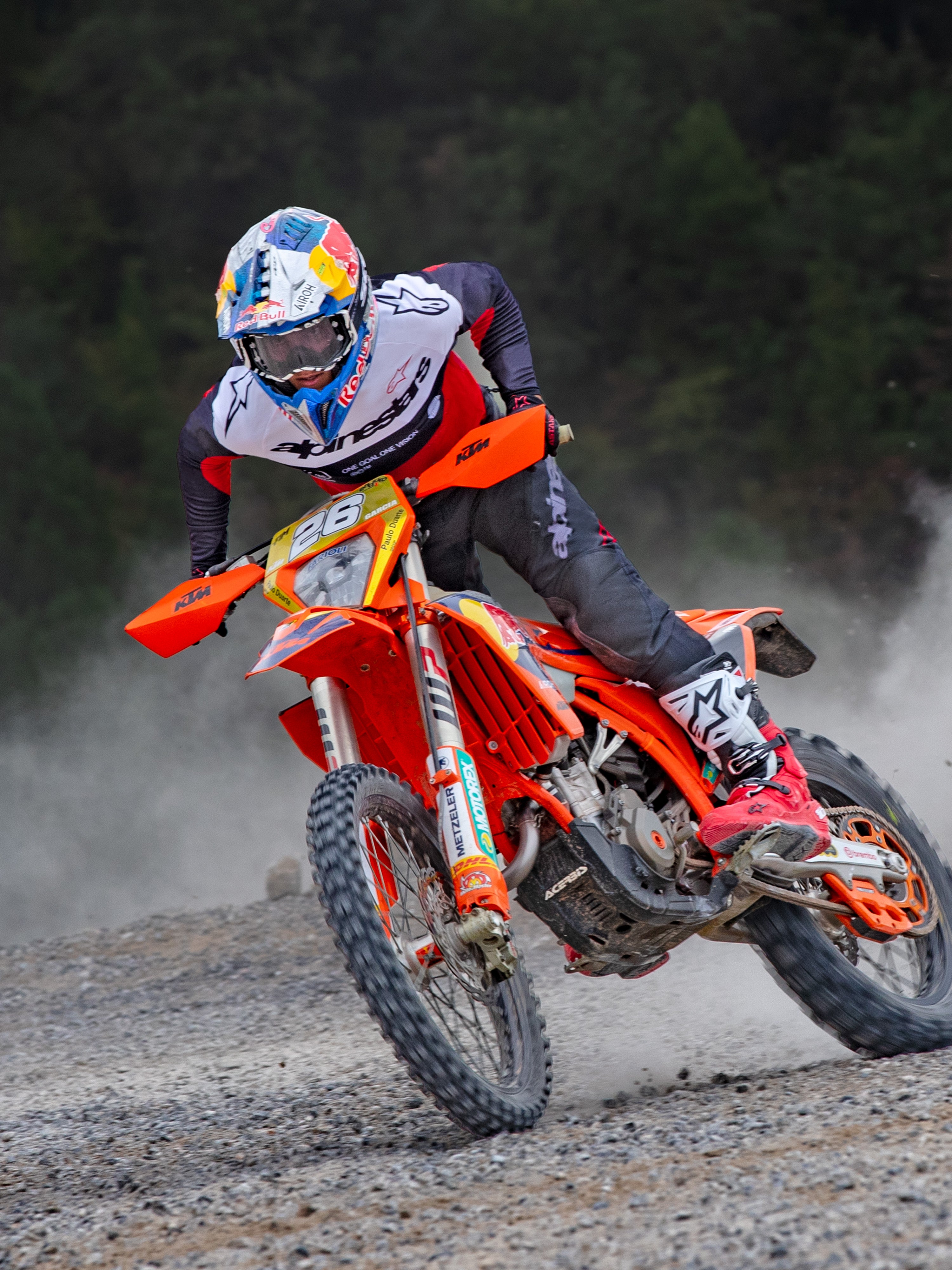Motocrossförare i action bärande Alpinestars Pro-Dura Jersey i svart, klarröd och vit, körandes en orange motocrosscykel på ett dammigt terrängspår, kombinerat med svarta byxor och röd-vita stövlar.