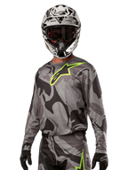 Alpinestars 2024 Racer Tactical Jersey, motocrosströja, grått kamouflagemönster med neongrön logotyp, bärs med svartvit hjälm och svarta handskar, tekniskt fukttransporterande material för terrängkörning