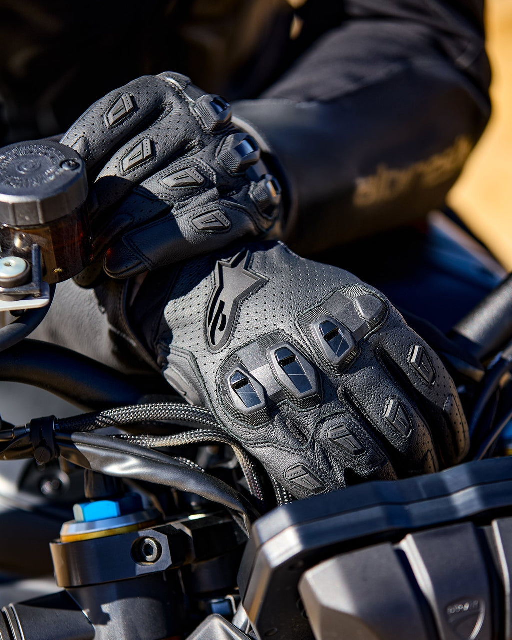 Alpinestars SP X 7-handskar, svarta motorcykelhandskar i perforerat läder med TPU-knogskydd och fingersliders, visas på en förares händer som greppar ett motorcykelstyre