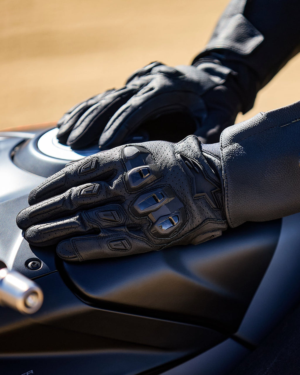 Alpinestars SP X 7 Handskar, svarta korta motorcykelhandskar i perforerat läder, händer vilande på en mörk bränsletank, med synligt TPU-knogskydd med dubbel densitet och präglad logotyp, designade för sport- och landsvägskörning