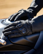 Alpinestars SP X 7 Handskar, svarta korta motorcykelhandskar i perforerat läder, händer vilande på en mörk bränsletank, med synligt TPU-knogskydd med dubbel densitet och präglad logotyp, designade för sport- och landsvägskörning