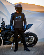 Alpinestars T-Stunt Air-jacka, sportigt skuren textiljacka i svart, mörkblå och ljusblå, bärs av en förare som står bredvid en Ducati-motorcykel, utrustad med kolfiberhjälm och svart körutrustning i ett ökenlandskap vid solnedgången.