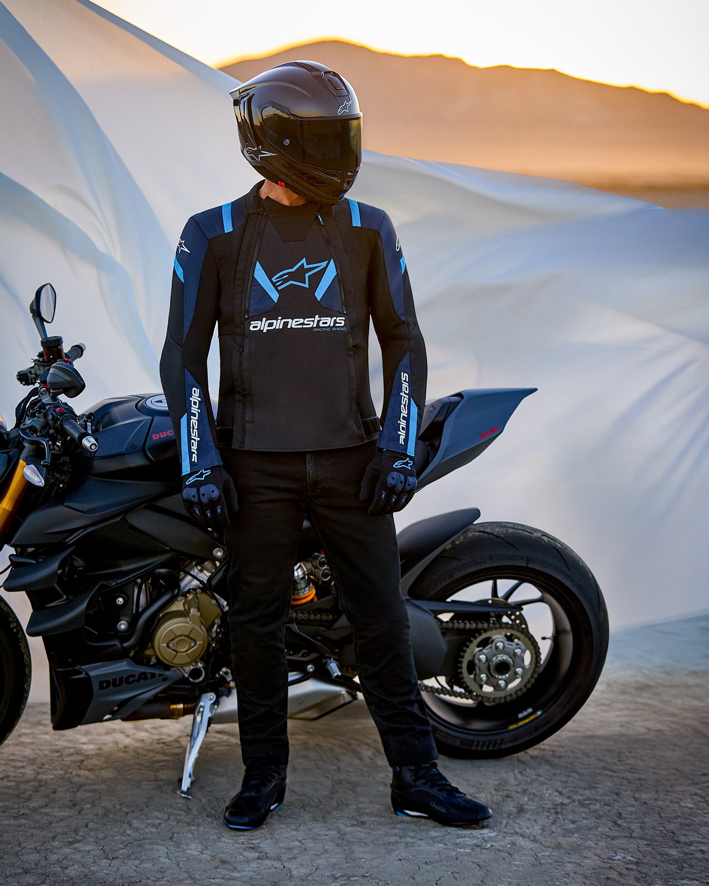Alpinestars T-Stunt Air-jacka, sportigt skuren textiljacka i svart, mörkblå och ljusblå, bärs av en förare som står bredvid en Ducati-motorcykel, utrustad med kolfiberhjälm och svart körutrustning i ett ökenlandskap vid solnedgången.