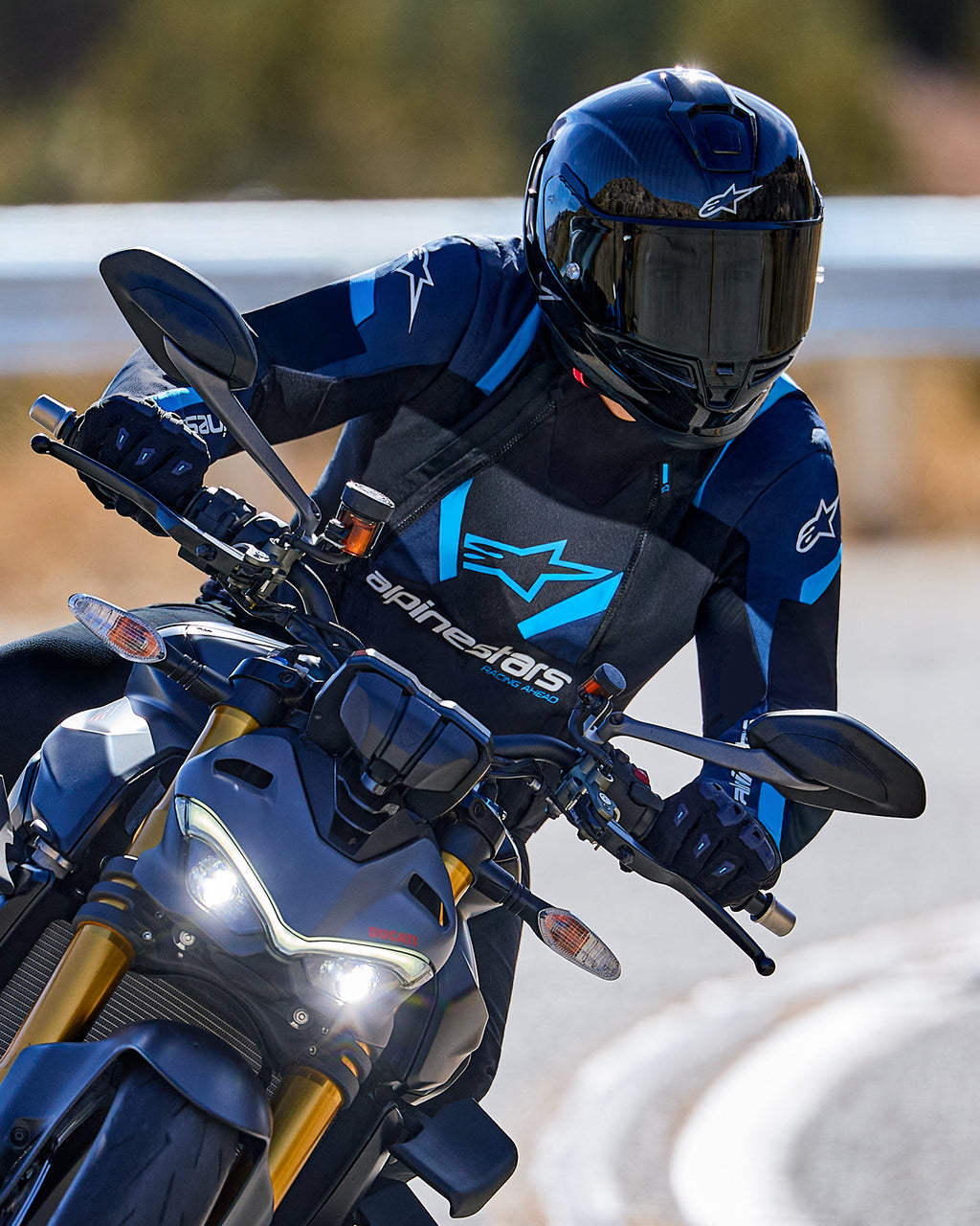 Motorcyklist som lutar sig in i en kurva iklädd Alpinestars T-Stunt Air-jacka i svart, mörkblått och klarblått, tillsammans med svart hjälm och handskar, actionbild på asfalterad väg, sportig körutrustning