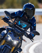 Motorcyklist som lutar sig in i en kurva iklädd Alpinestars T-Stunt Air-jacka i svart, mörkblått och klarblått, tillsammans med svart hjälm och handskar, actionbild på asfalterad väg, sportig körutrustning