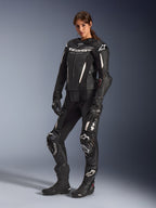 Kvinna bär Alpinestars Stella GP-R7 2-delat skinnställ i svart, professionell racingutrustning för motorcykel med vita logotypdetaljer och accenter, matchat med svarta racinghandskar och stövlar, helkroppsbild i studio.