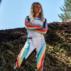 Female motocross rider wearing Alpinestars Women Stella 2024 Techstar Pants and matching jersey, Black Multicolor design with white base and vibrant blue, yellow, and red stripes, standing with arms crossed on a dirt track, professional off-road racing outfit