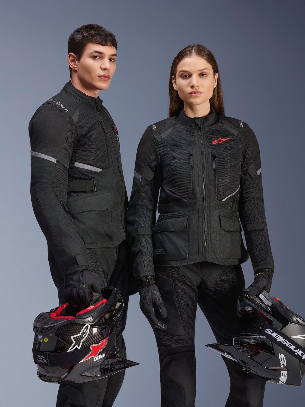 Manlig och kvinnlig modell iklädda Alpinestars Stella Andes Air Drystar®-jackor i svart, adventure-touringutrustning för motorcykel med röda stjärnlogotyper, ventilationspaneler i mesh och teknisk textilkonstruktion, modeller som håller i adventure-hjälmar i en studiobild