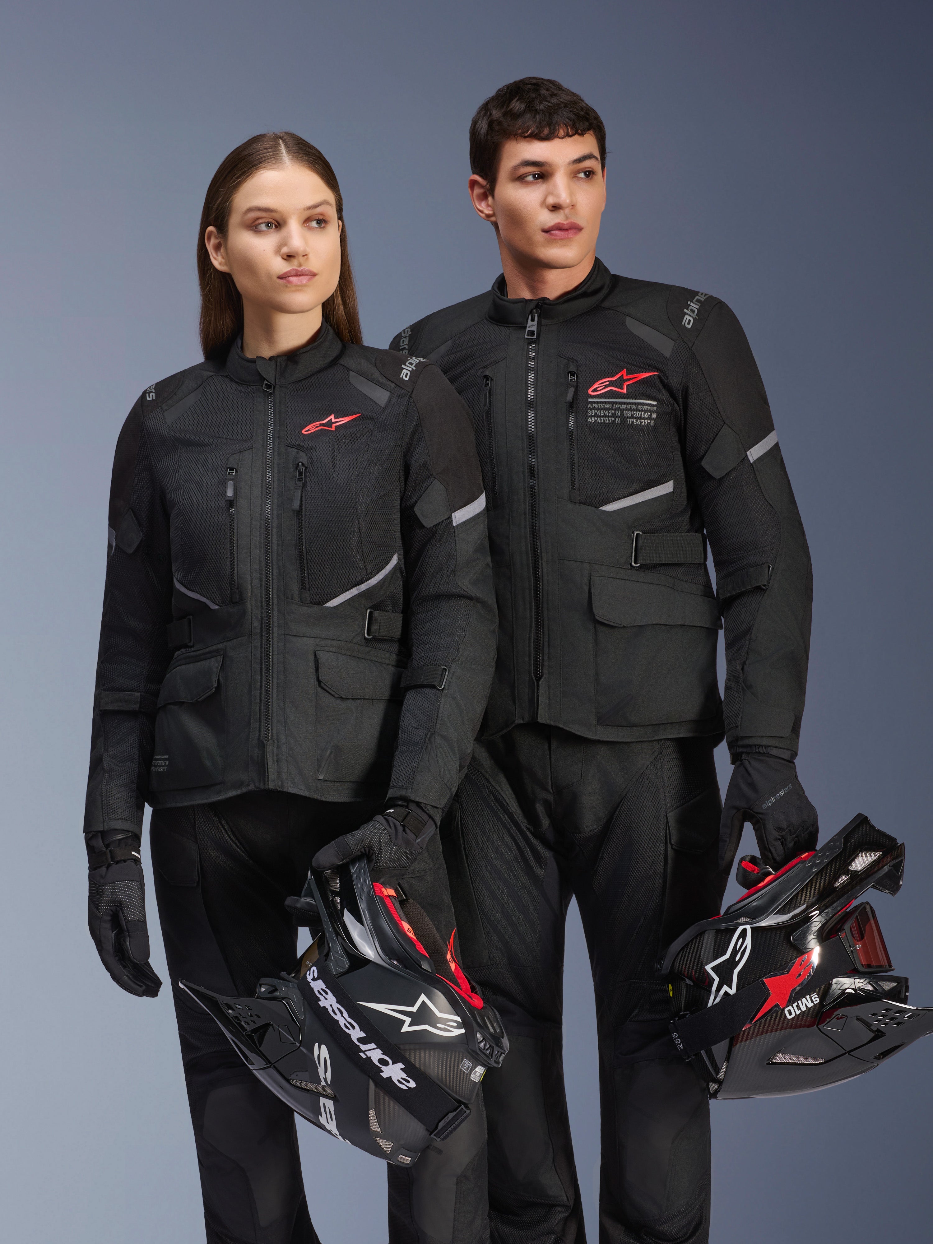 Man and woman wearing Alpinestars Andes Air Drystar® Jackets, Black adventure touring gear with mesh panels and reflective details, matching black pants and gloves, holding motocross helmets, studio shot of professional motorcycle apparel